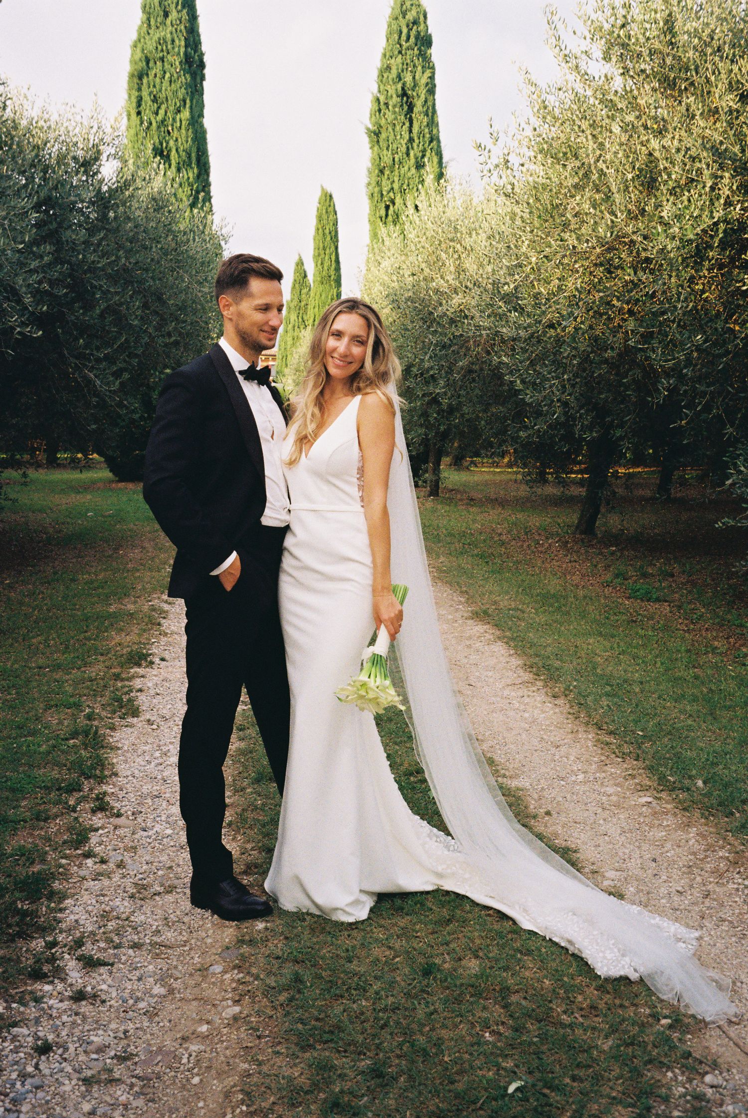 A wedding couple poses on a path lined with cypress trees and olive groves in Italy.