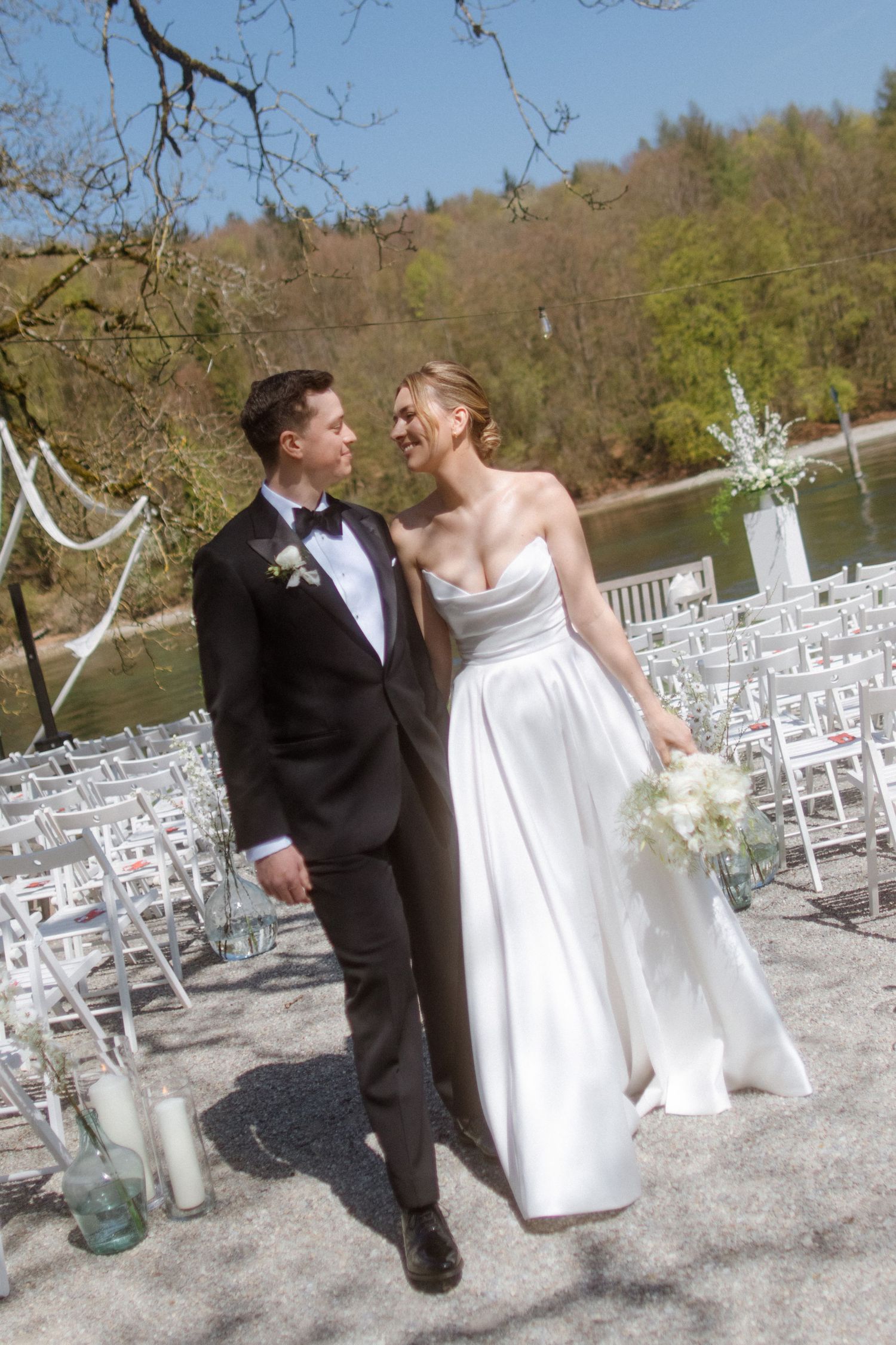 A smiling bride and groom walk together down the aisle at a lakeside outdoor wedding ceremony on a sunny day.