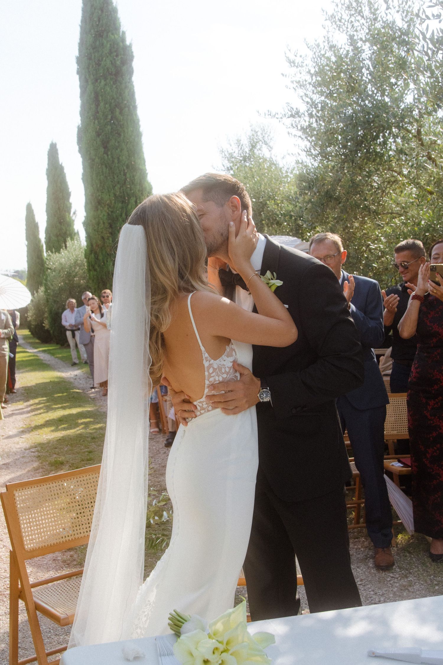 Newlyweds share their first kiss during outdoor ceremony with cypress trees backdrop.
