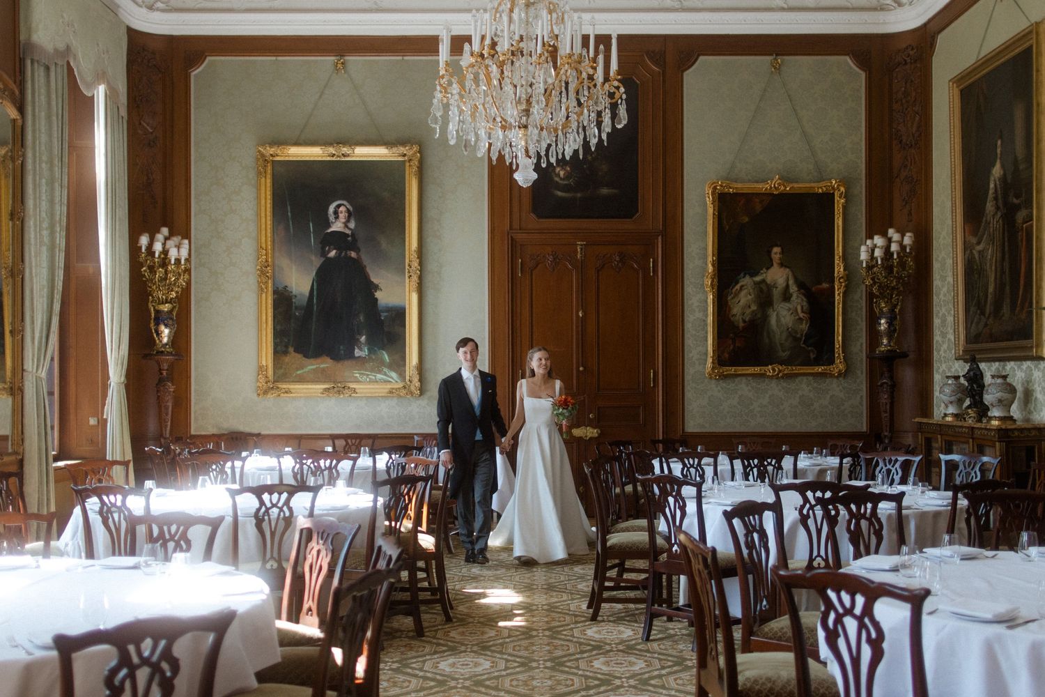 A wedding couple stands in an elegant dining room with crystal chandeliers and portrait paintings.