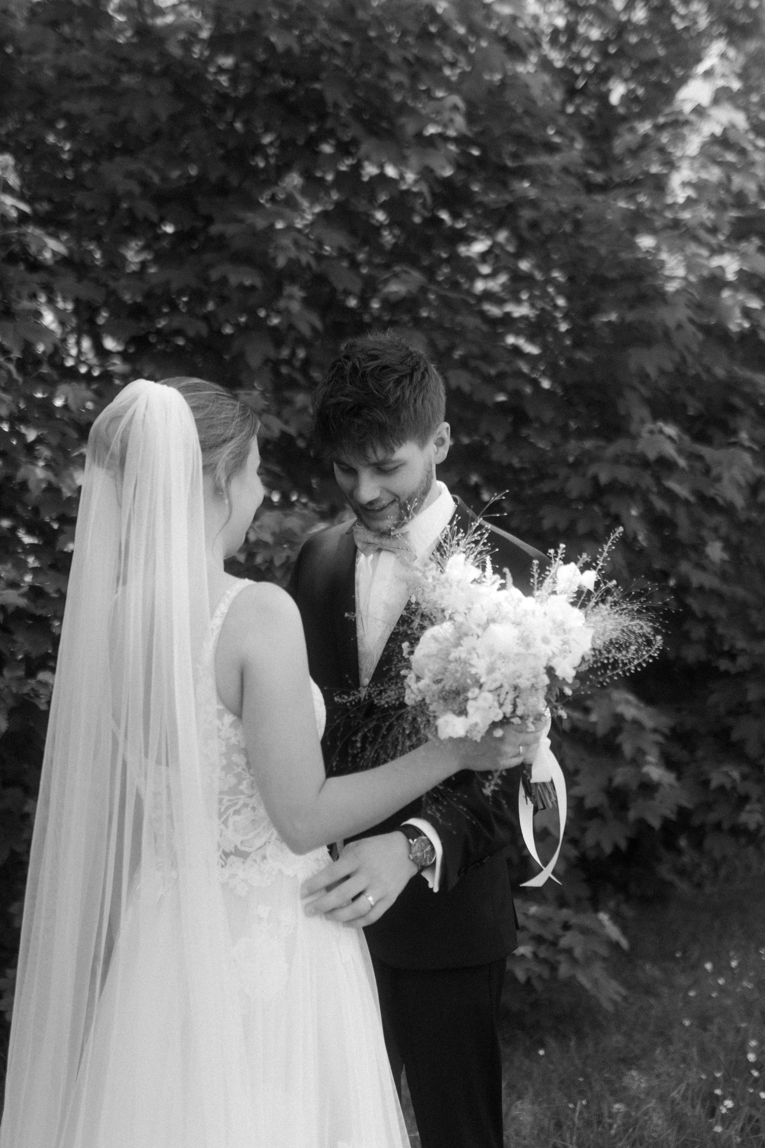 Black and white photo of newlyweds sharing a tender moment with a white bouquet in a garden.