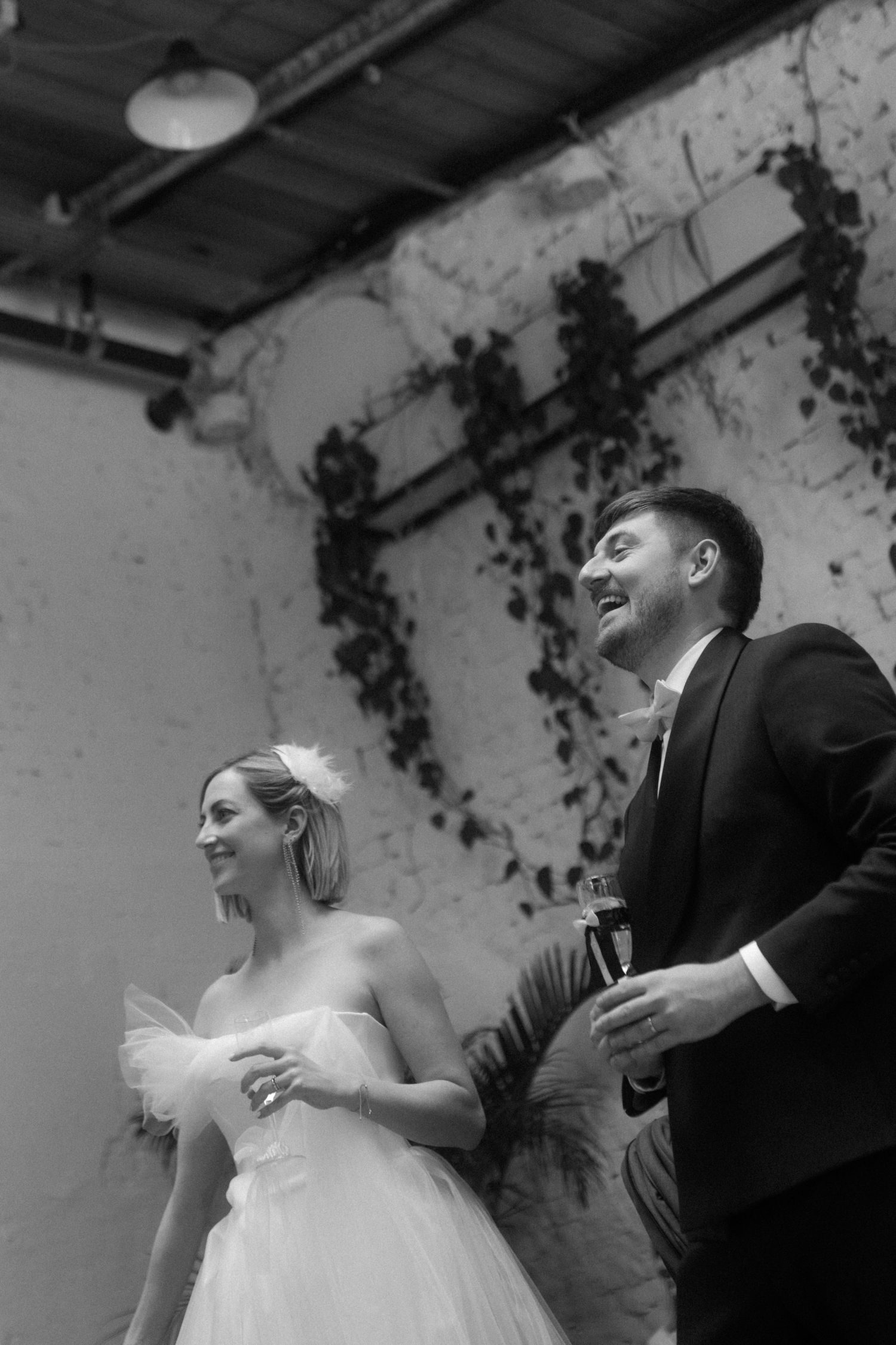 A bride in a white gown and groom in a suit smile while standing together in an industrial venue with hanging vines.
