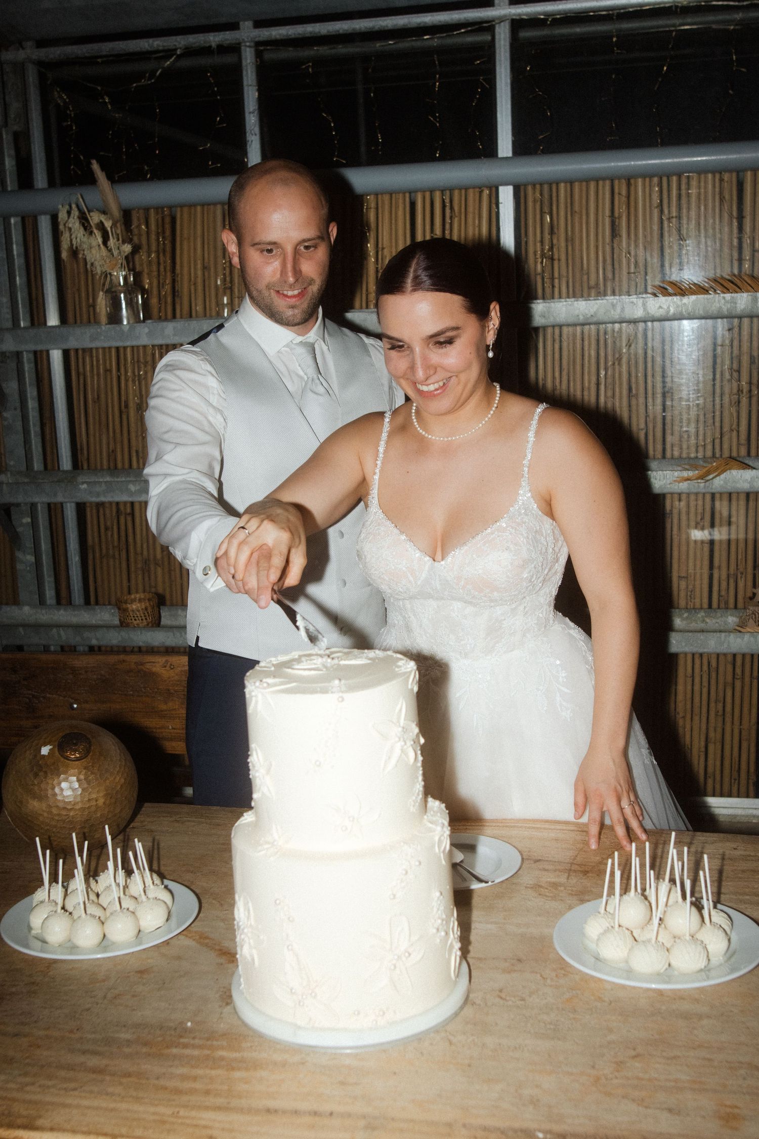 A couple cuts their elegant white wedding cake together while surrounded by cake pops on wooden tables.