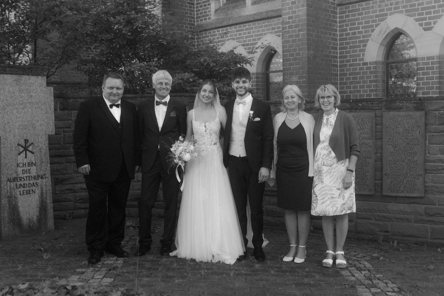 Small wedding party group poses for formal photo against brick church exterior.