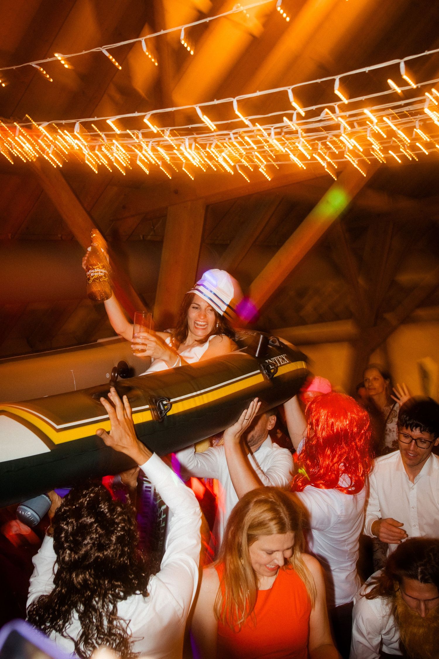 Party crowd carries person on inflatable raft under string lights at celebration.