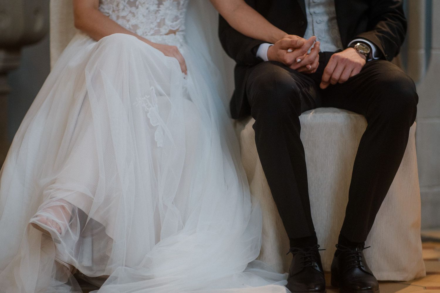 A close-up view of a bride's flowing white dress and groom's formal attire as they sit together.