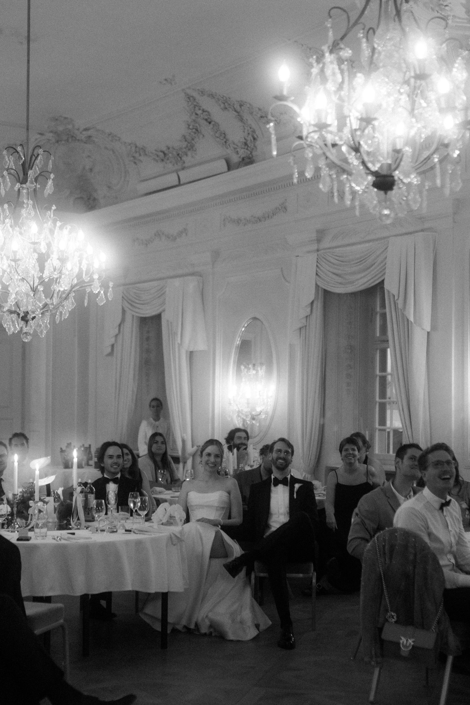 Wedding celebration in a luxurious white ballroom with crystal chandeliers and draped windows.