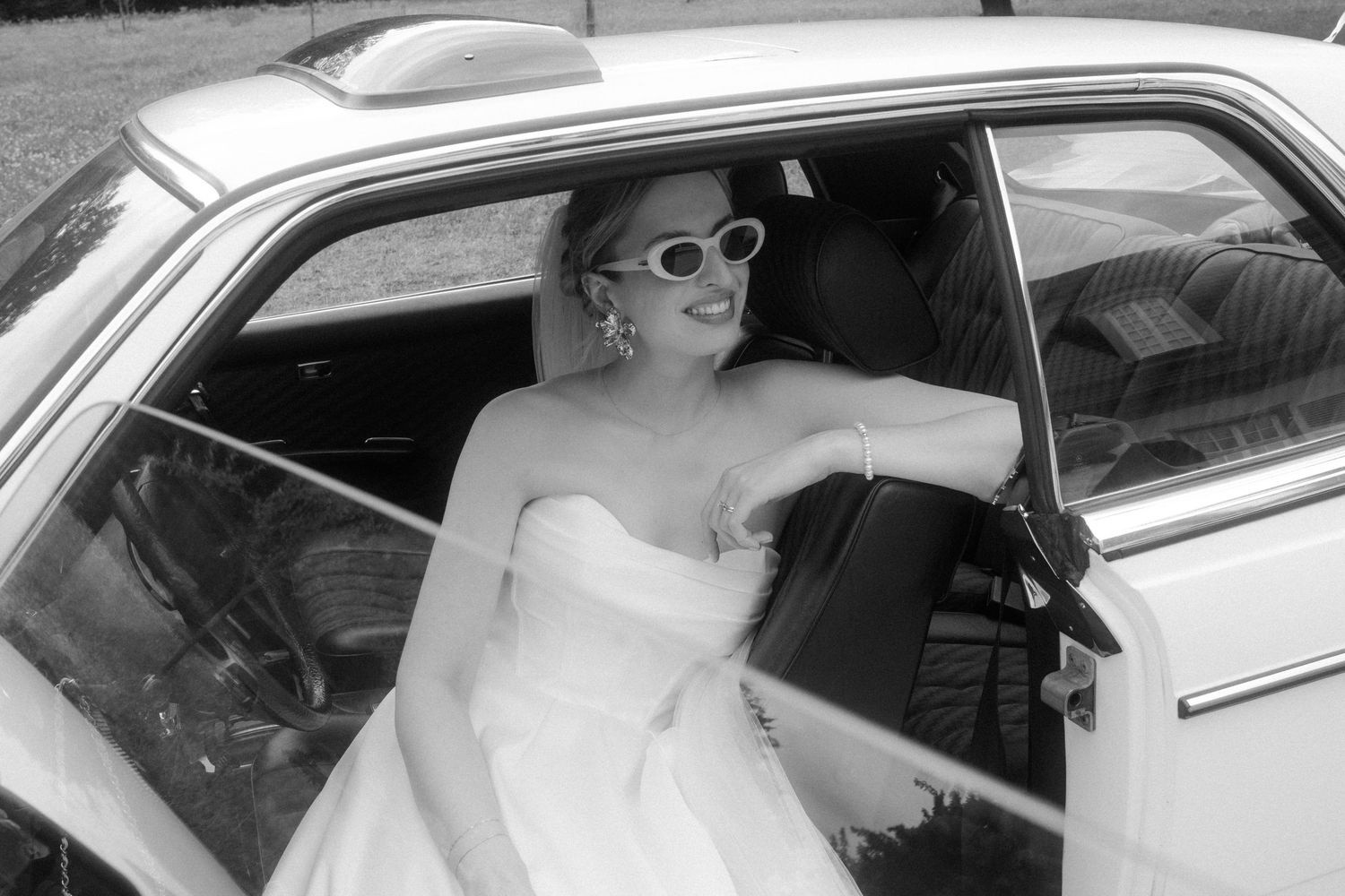 Bride sits in vintage car while groom stands outside during wedding photoshoot.