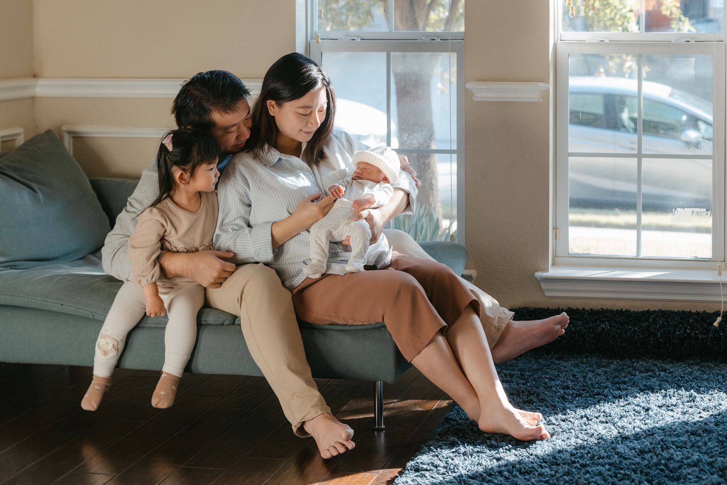 A couple sits comfortably on a couch by a window with a young baby and small child in their casual living room setting.