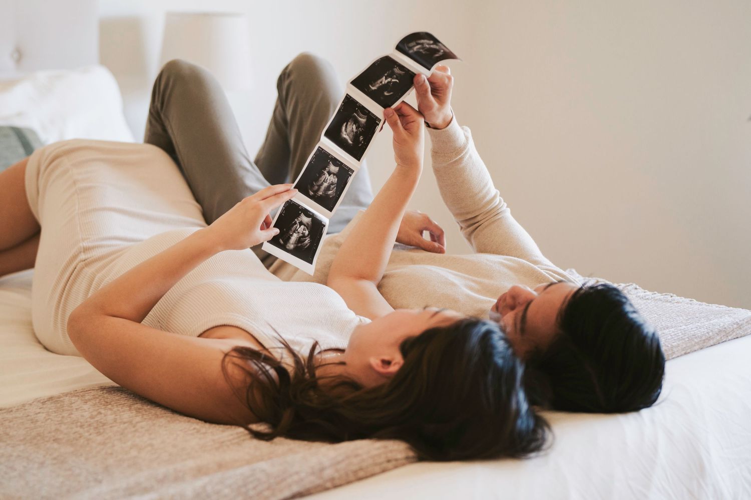 Person lying on white bed looking at strip of photo prints in casual relaxed pose.