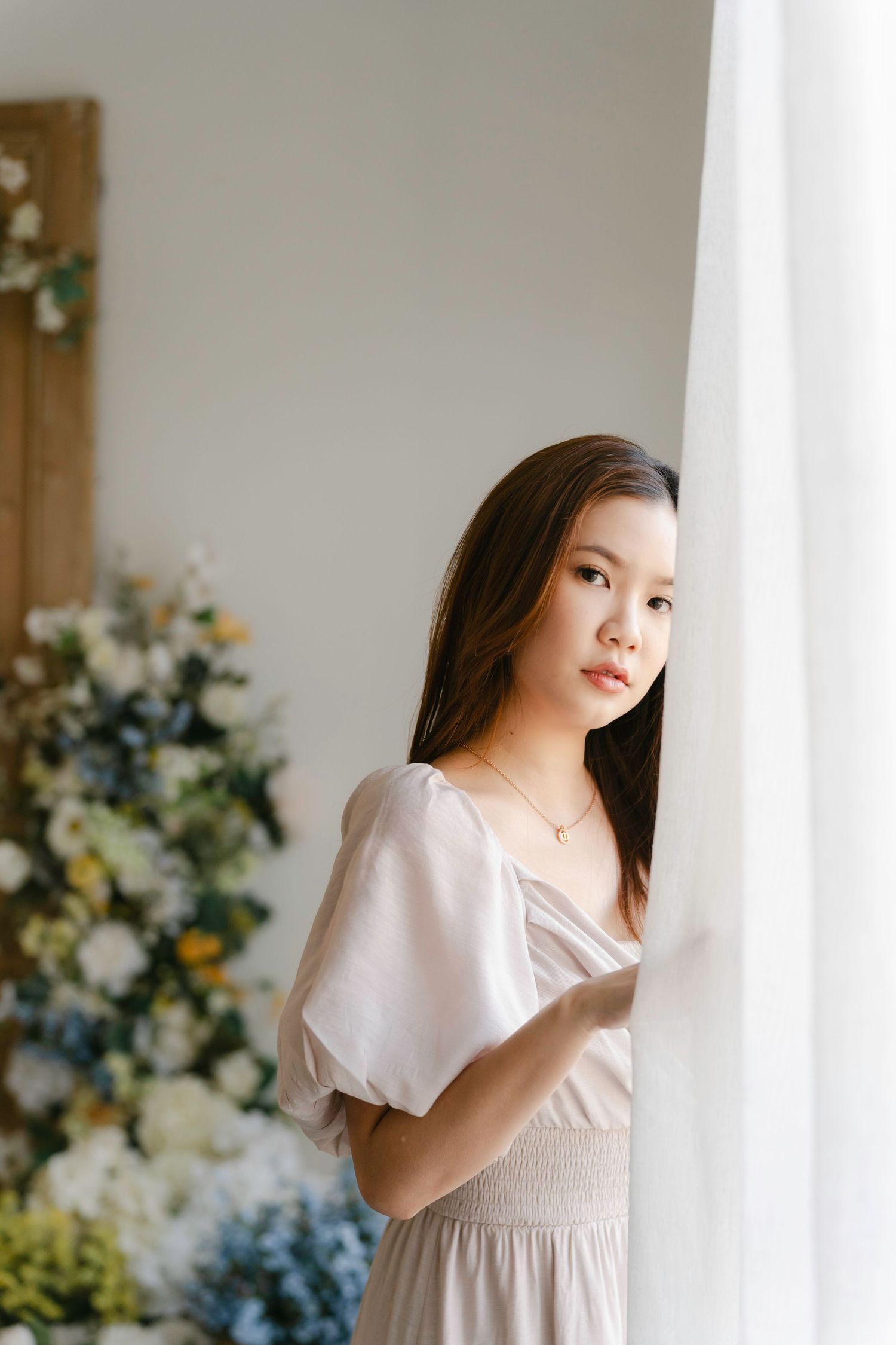 Figure in a light colored dress stands near curtained window with floral decorations in background.