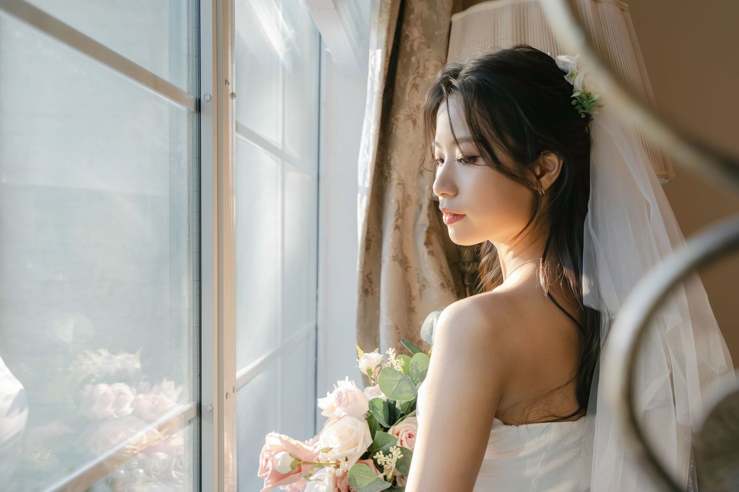 Bride in a white wedding dress gazes thoughtfully out a sunlit window while holding a delicate bouquet.