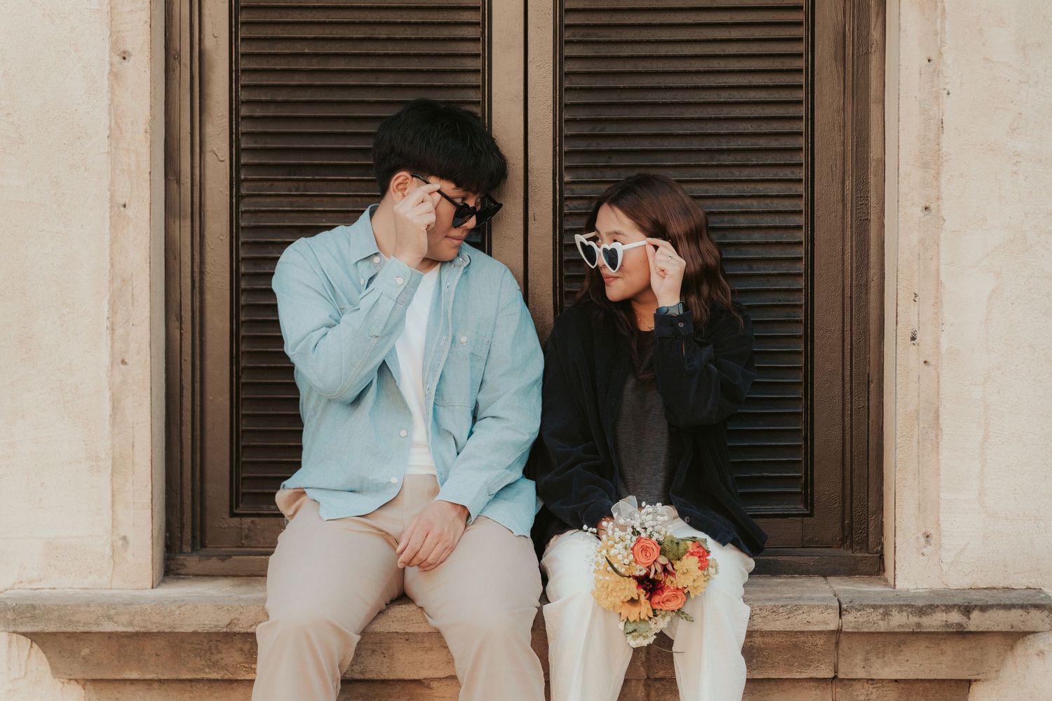 Happy couple in formal wear sits on stone ledge holding pink and orange flower bouquet during romantic photo session.