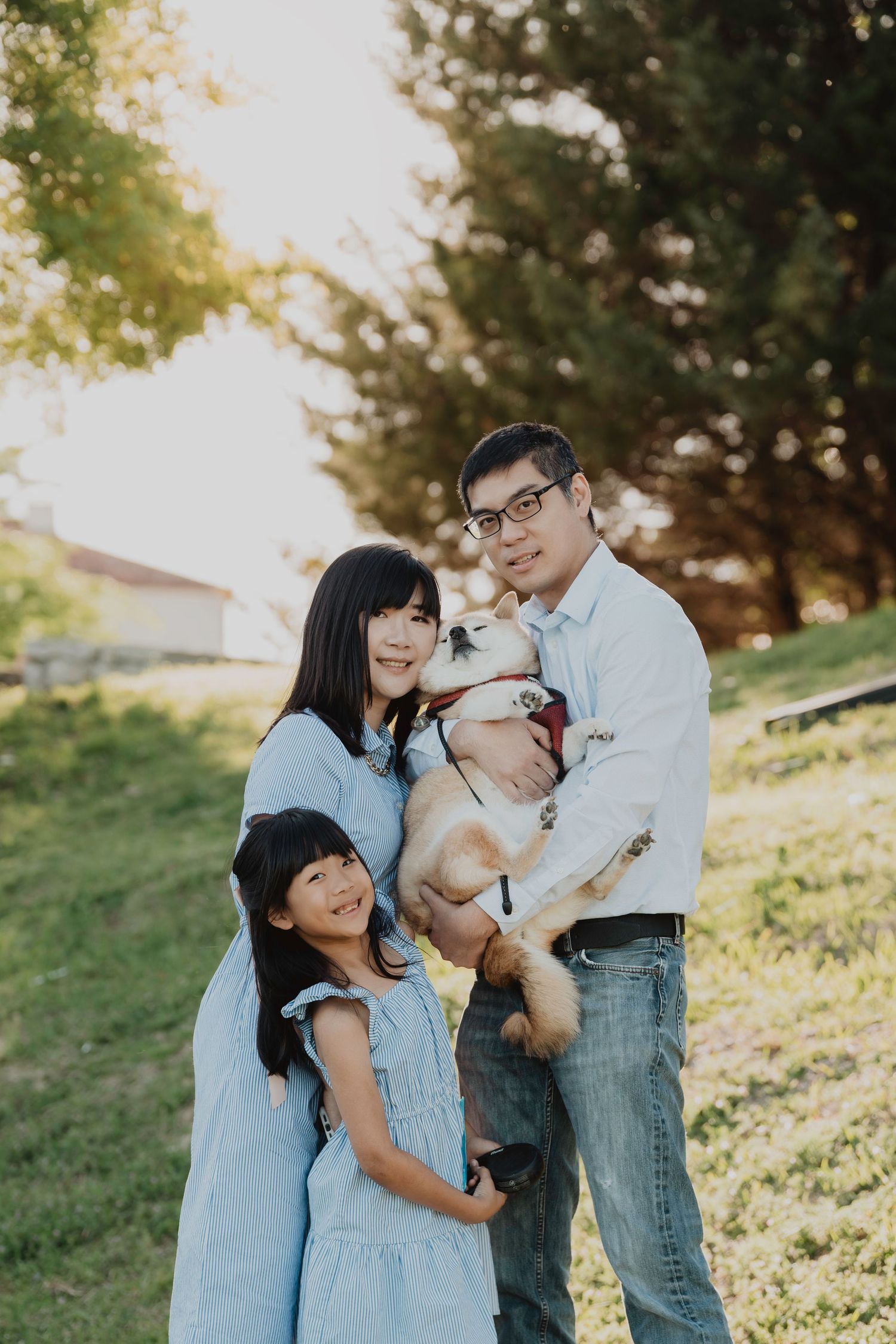 Family wearing coordinated blue outfits poses together outdoors with their small pug dog on a grassy hillside at sunset.
