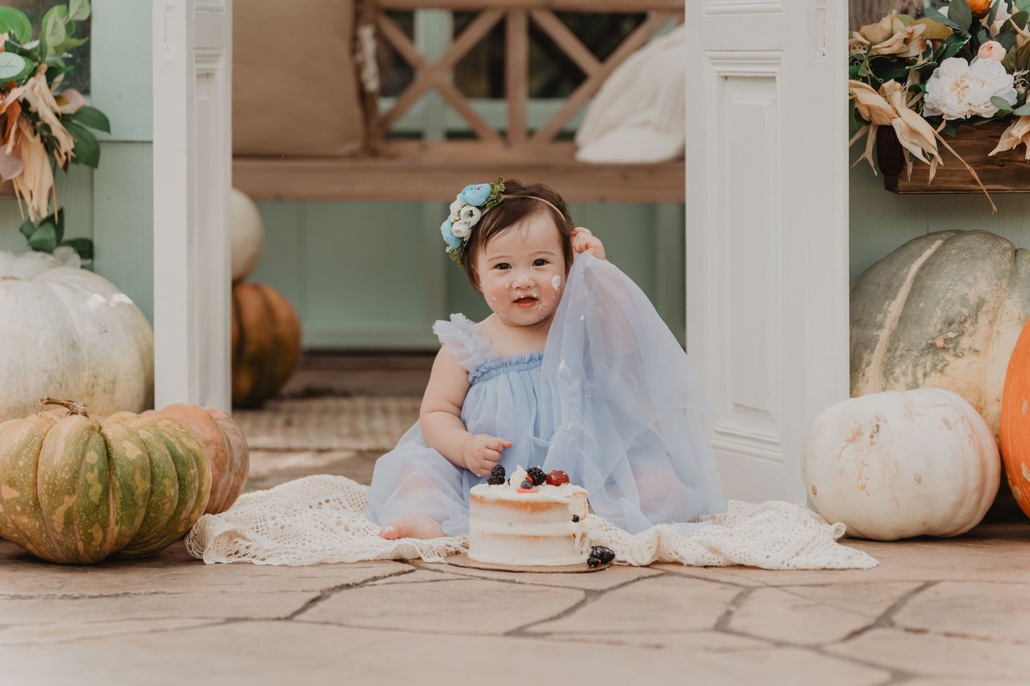 Small child in blue dress sits by white pumpkins on porch with a birthday cake and candles for cake smash photo.
