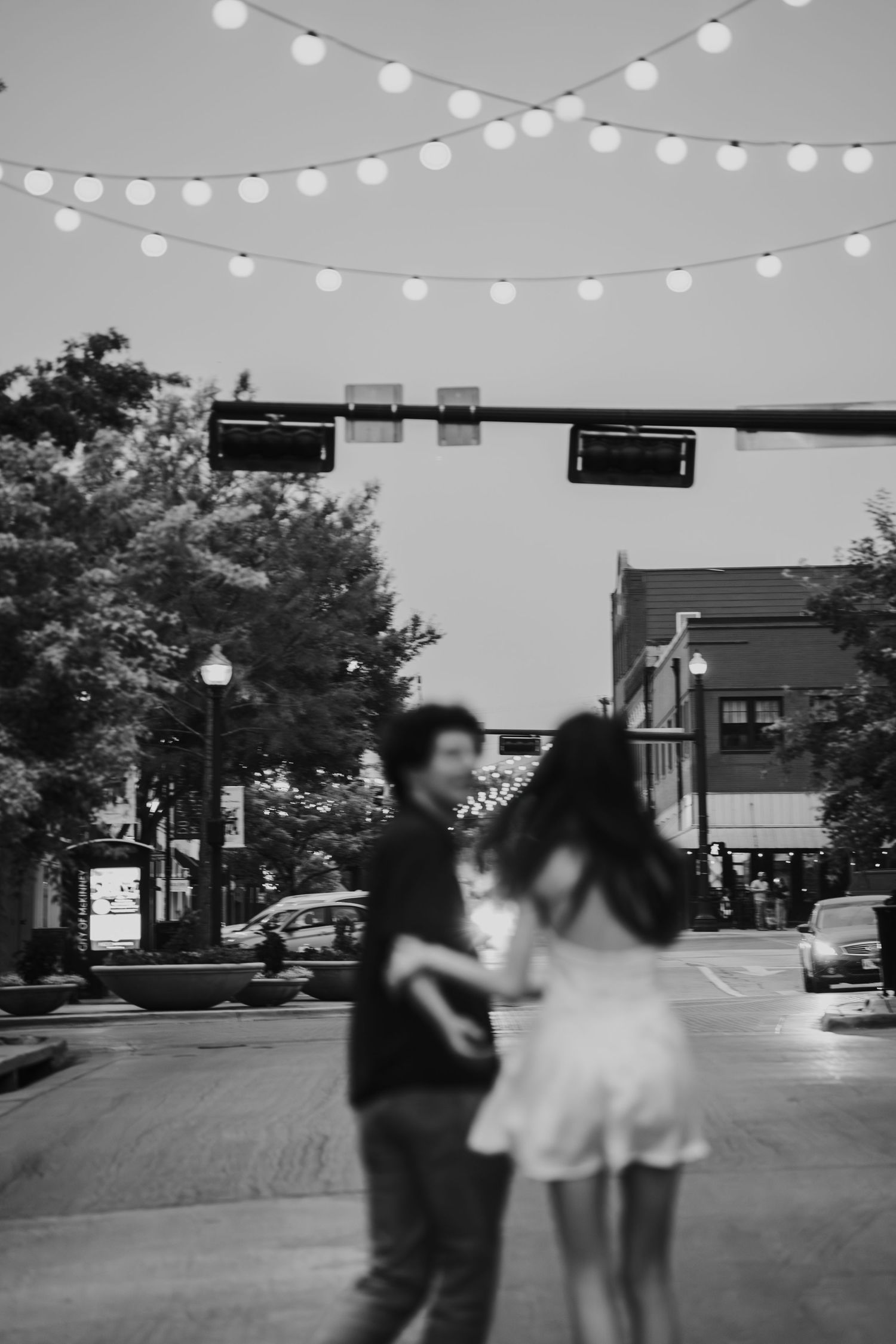 Black and white photo of a city street at dusk with string lights overhead and silhouettes dancing below.