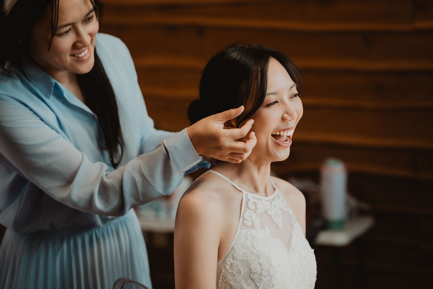 Bridesmaid in blue dress helping with wedding preparations as a joyful bride wearing lace gown gets ready.