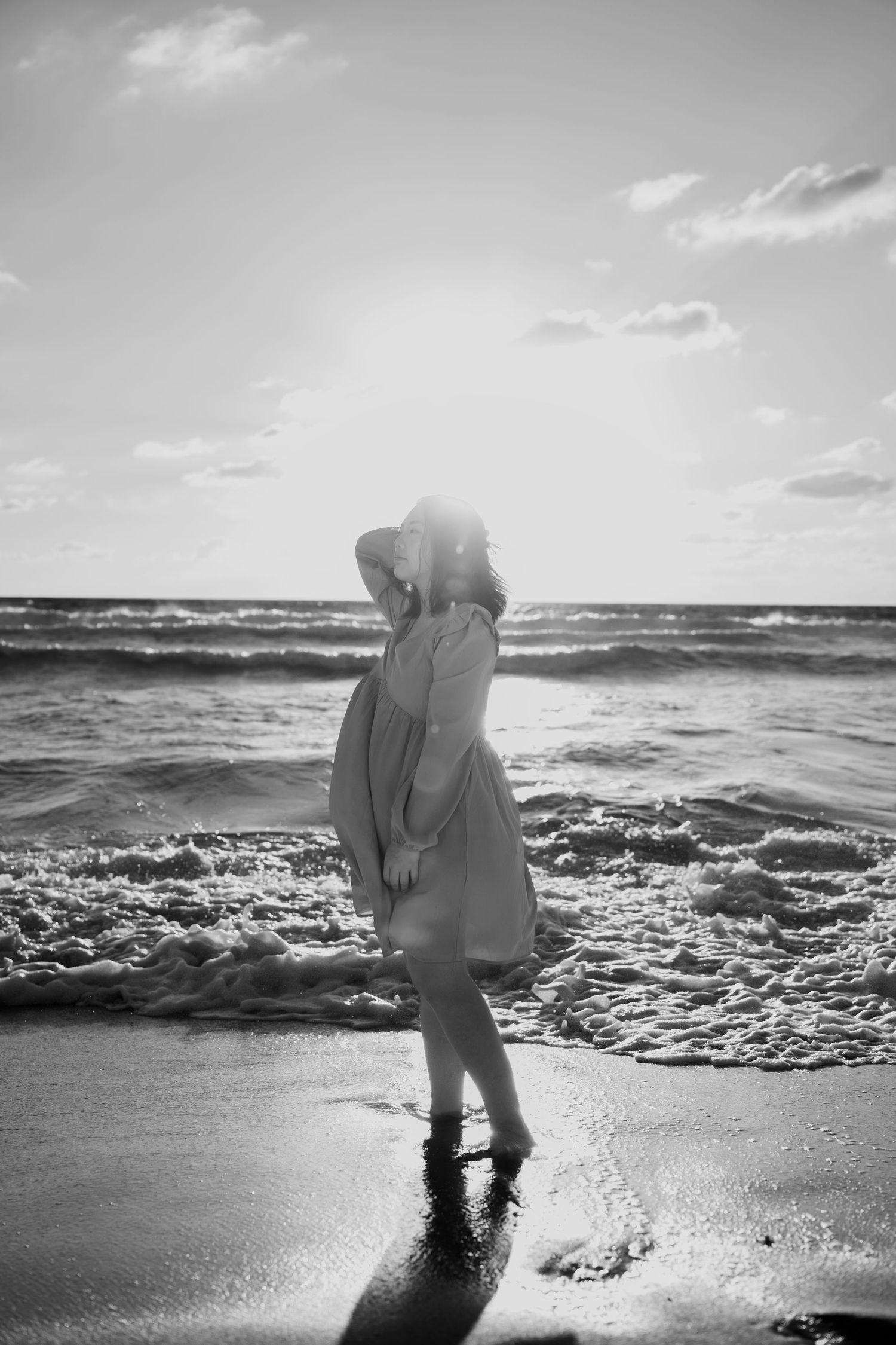 A silhouetted figure in a dress stands at the ocean's edge during sunset, waves crashing against the shore in black and white.