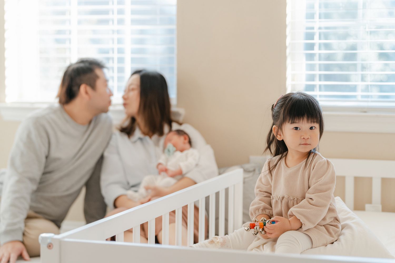 Young child in beige shirt sits on bed while parents share intimate moment with newborn baby in background.