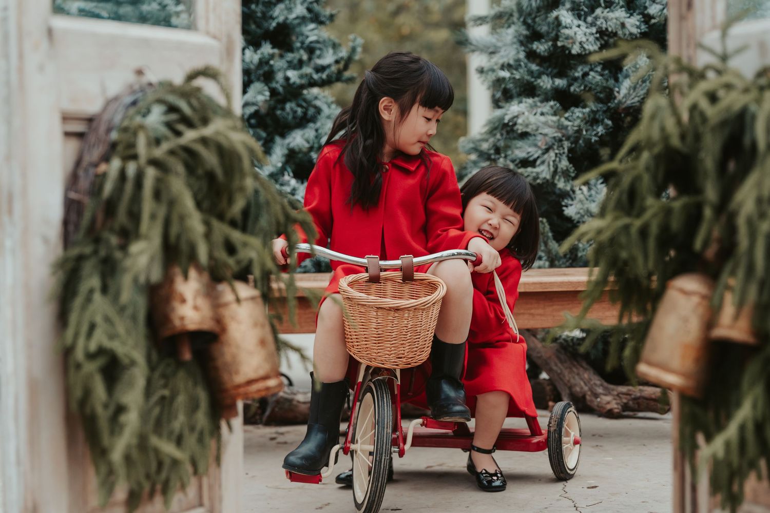 Two children in matching red outfits play together with a vintage bicycle and tricycle between potted evergreen plants.