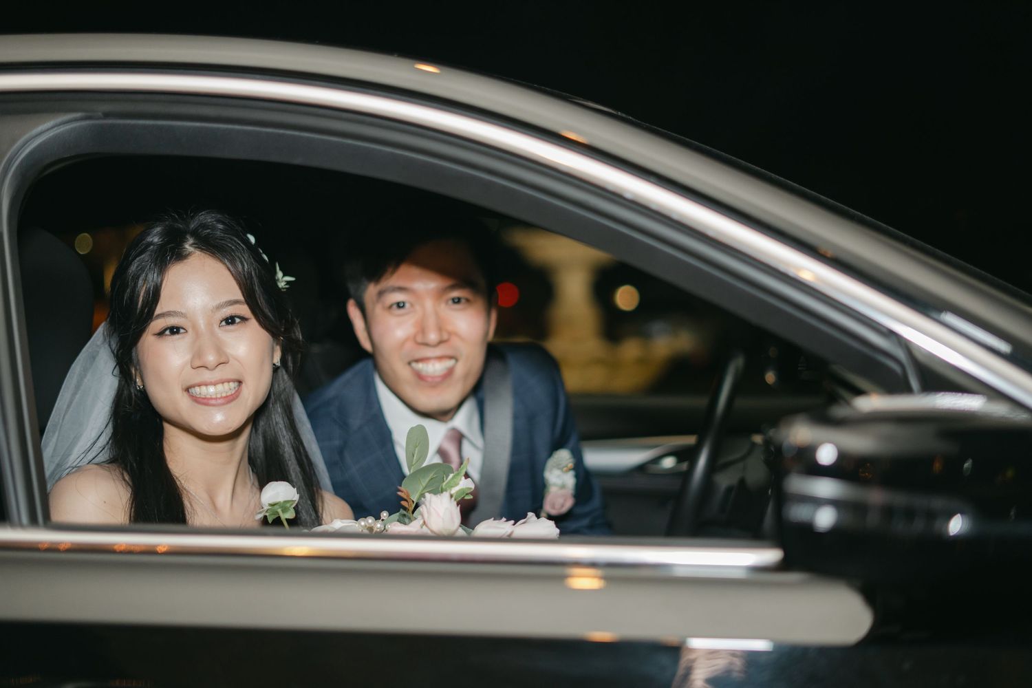 Happy bride and groom smile from inside their car at a wedding venue during the evening.
