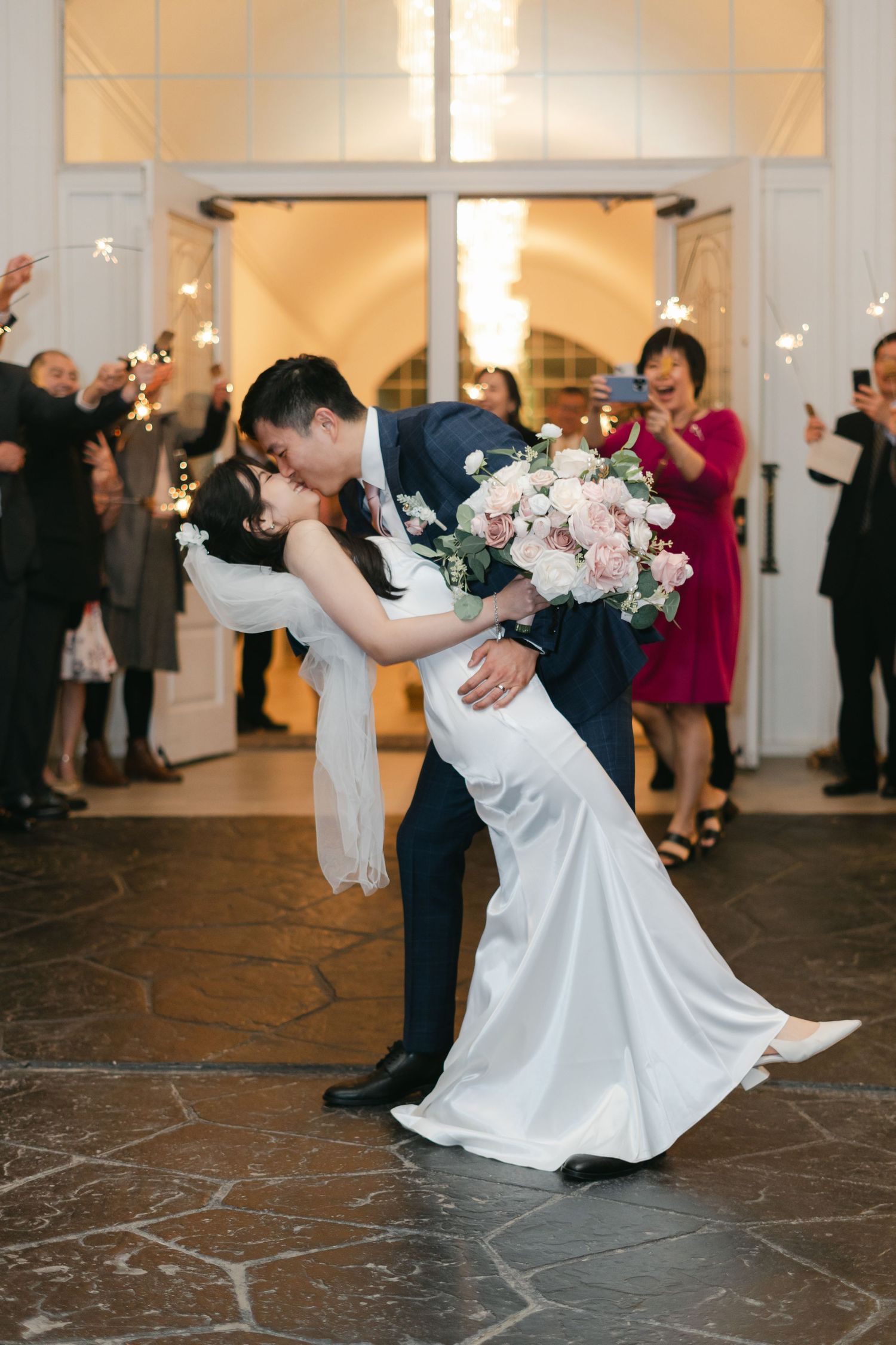 Newlyweds share romantic dip kiss during first dance while wedding guests cheer with sparklers in elegant venue.
