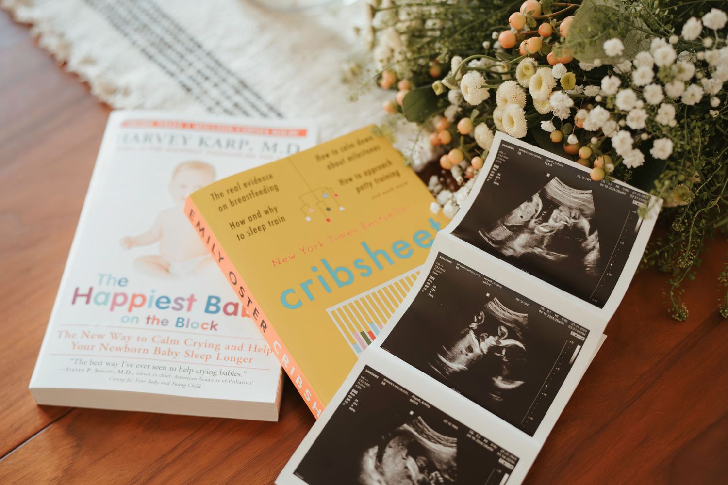 Pregnancy ultrasound photos displayed alongside parenting books and baby's breath flowers on wooden surface.