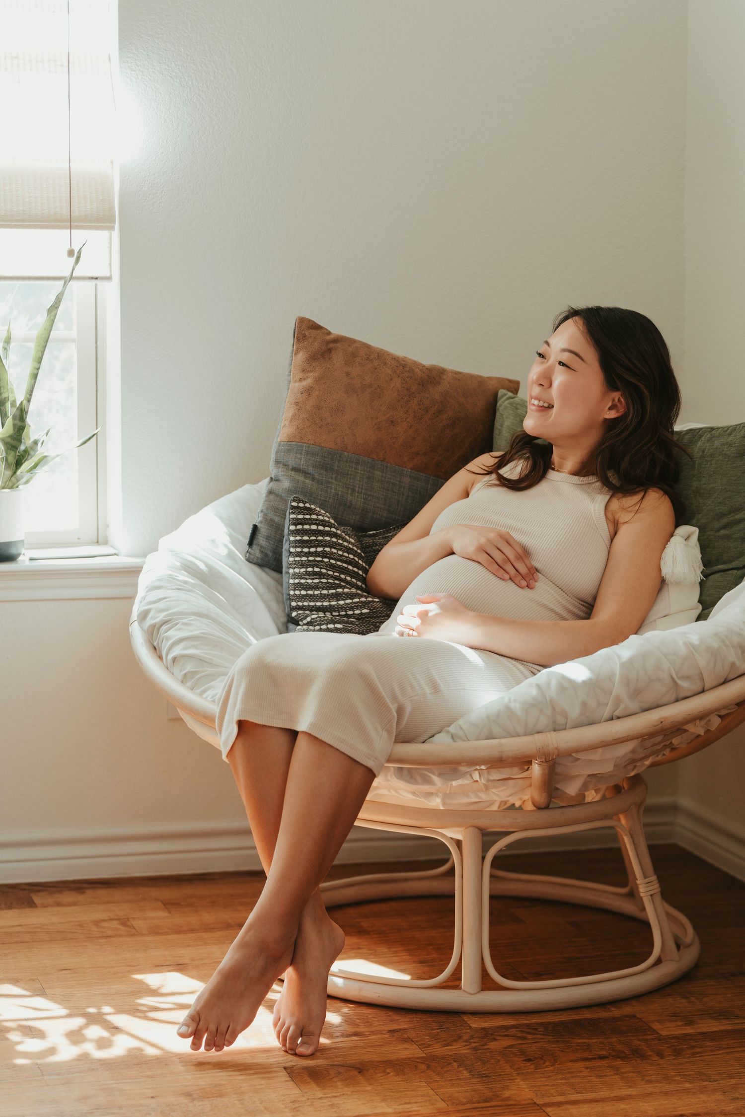 Woman relaxes in cozy rattan papasan chair by window with decorative pillows and natural sunlight streaming in.