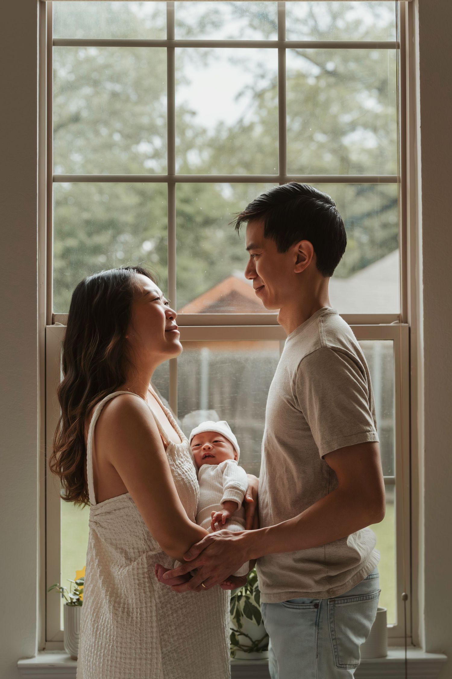 Young couple stands lovingly by a window holding their newborn baby in a tender intimate moment at home.