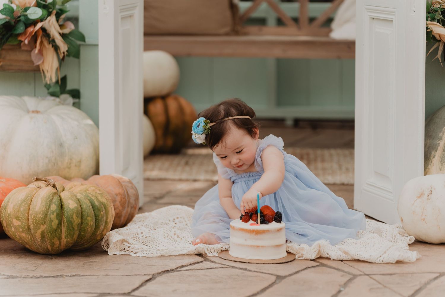 A small child in a blue dress sits on a porch with decorative pumpkins and a tiny cake during an autumn photoshoot.