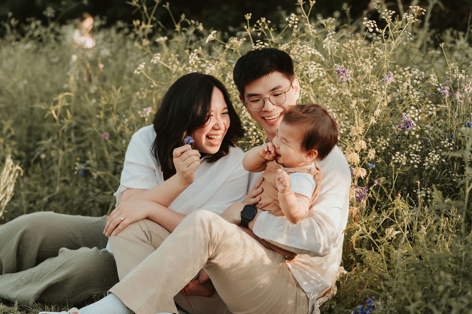 A family sits together laughing in a sunlit meadow with tall grass and wildflowers during golden hour.