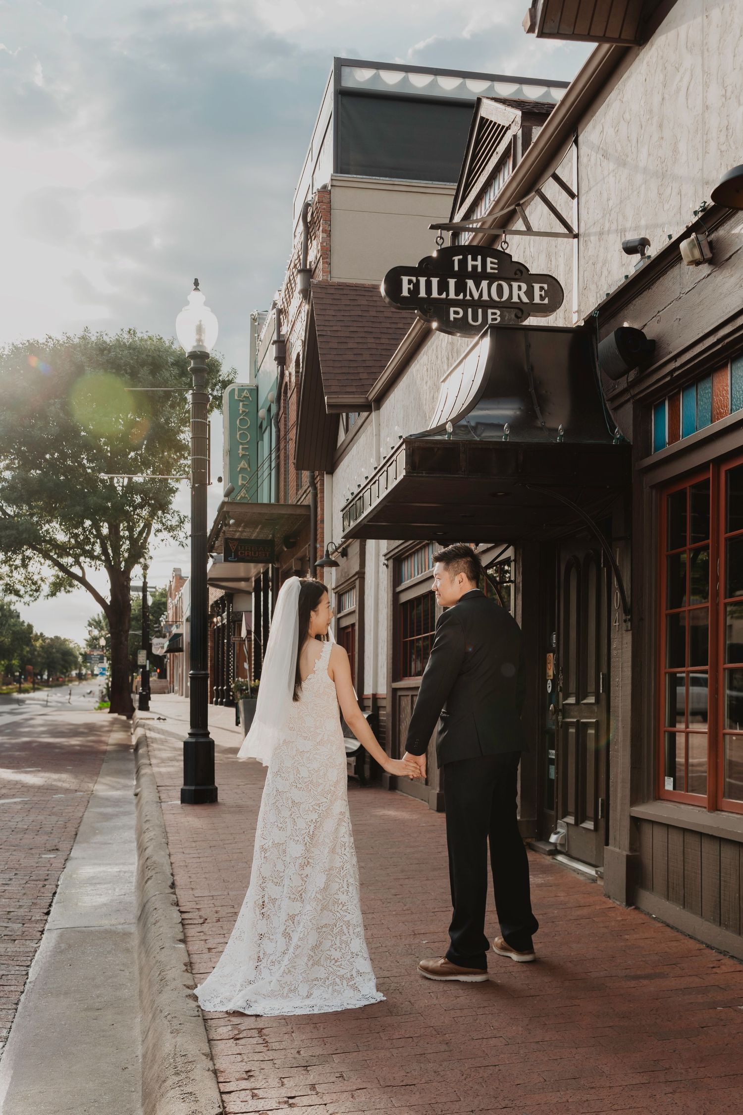 A couple in wedding attire holds hands while standing on a sidewalk beneath The Fillmore storefront sign.