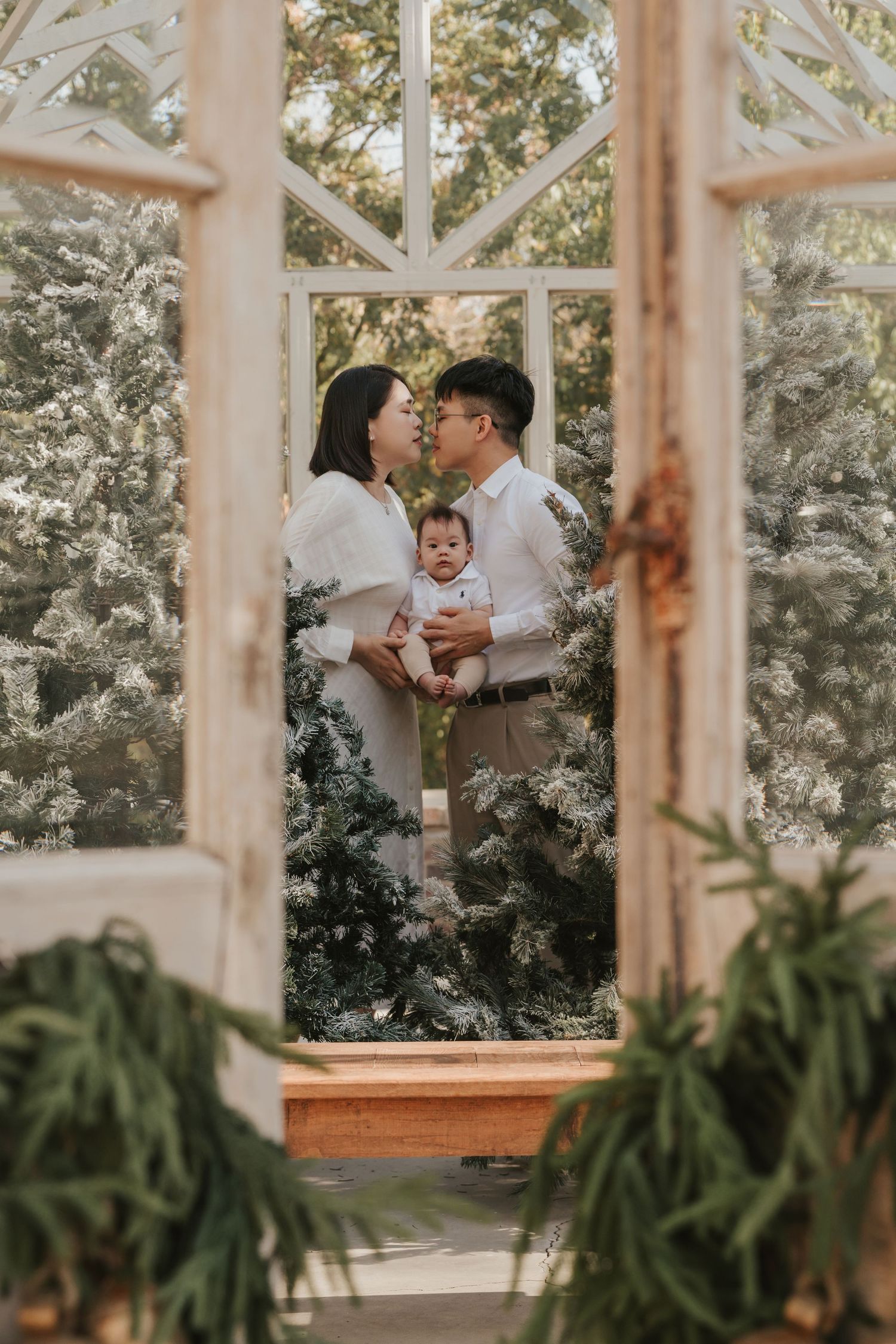 Family portrait viewed through glass doors of a greenhouse with lush foliage and natural light.