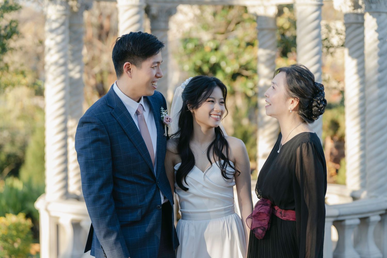 Three people share a joyful moment together outdoors in formal attire near elegant white columns on a sunny day.