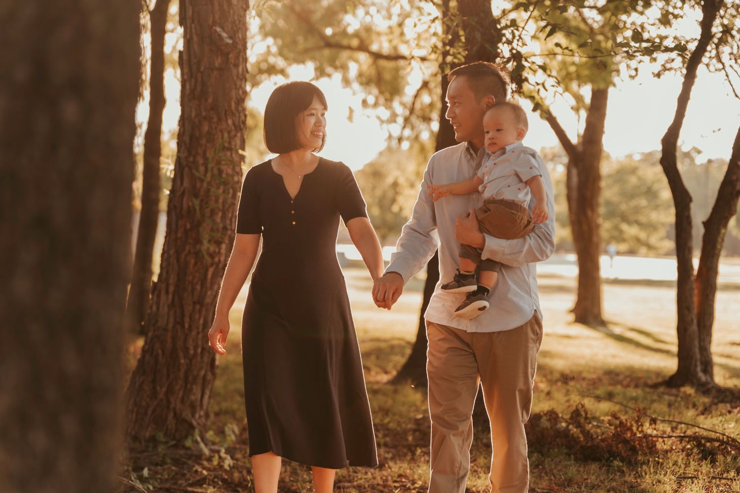 Couple holds hands while walking along wooded lakeside path during golden hour sunset.