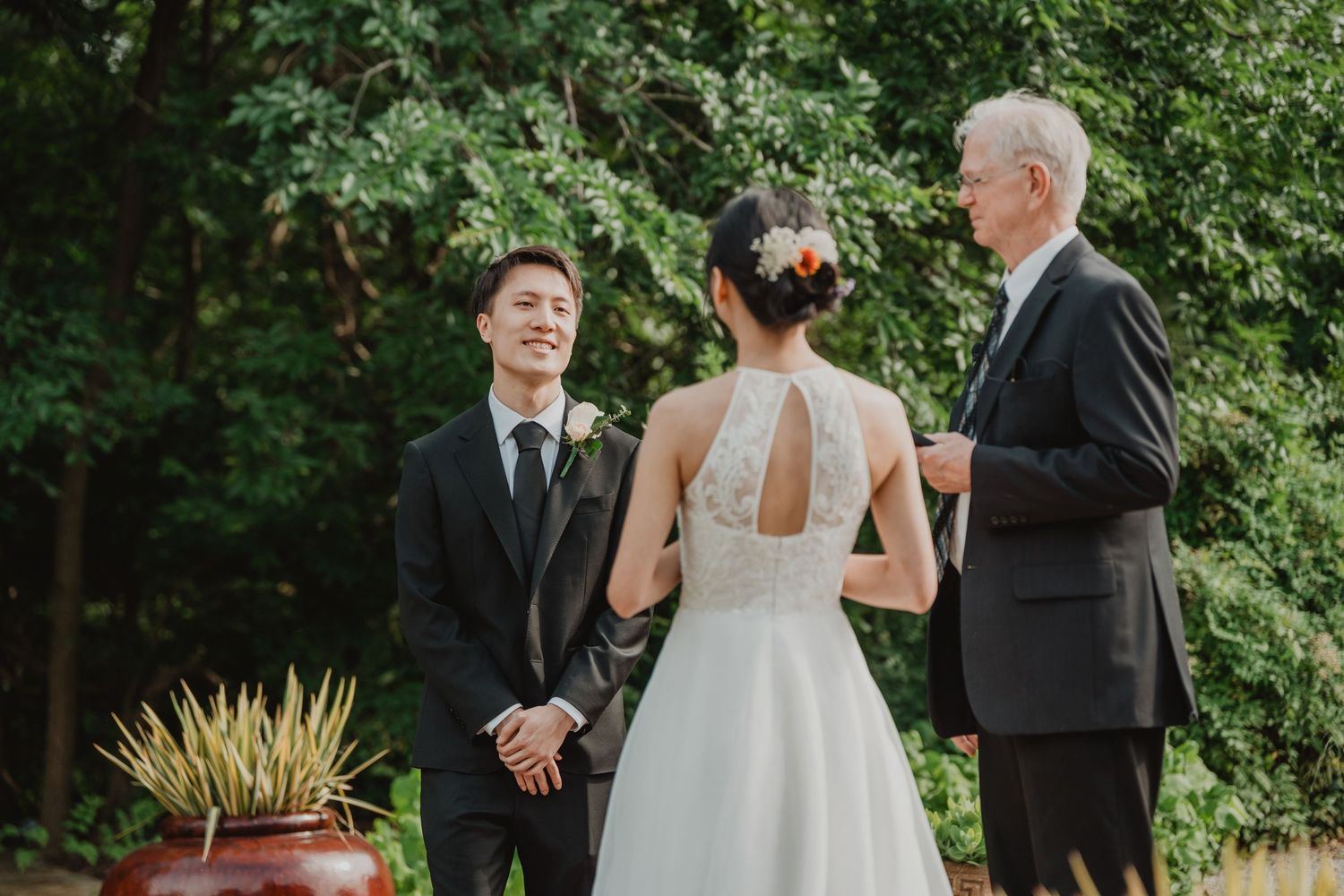 A wedding ceremony takes place outdoors with the bride in a white dress and two men in dark suits among green foliage.