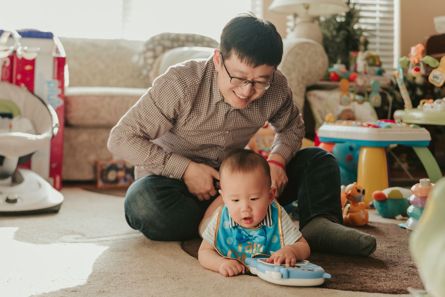 Adult and baby sitting on carpet looking at book together in living room with toys scattered around.