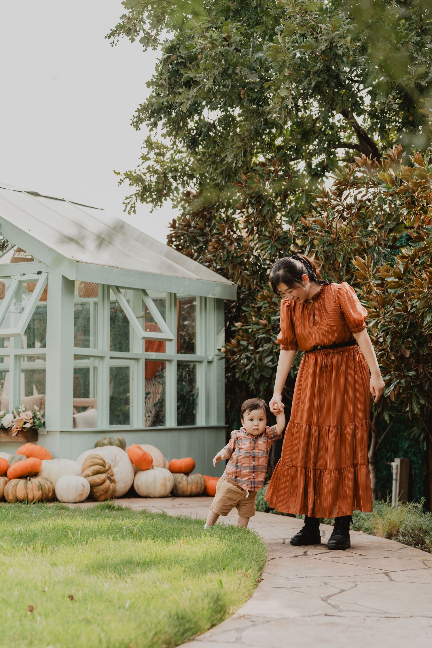 Fall scene with people in autumn-themed attire standing by greenhouse and pumpkin display in garden setting.