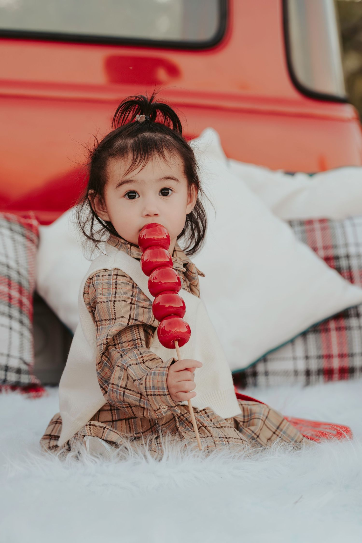A child in plaid dress holding red macarons on a skewer sits among white pillows with plaid pattern.