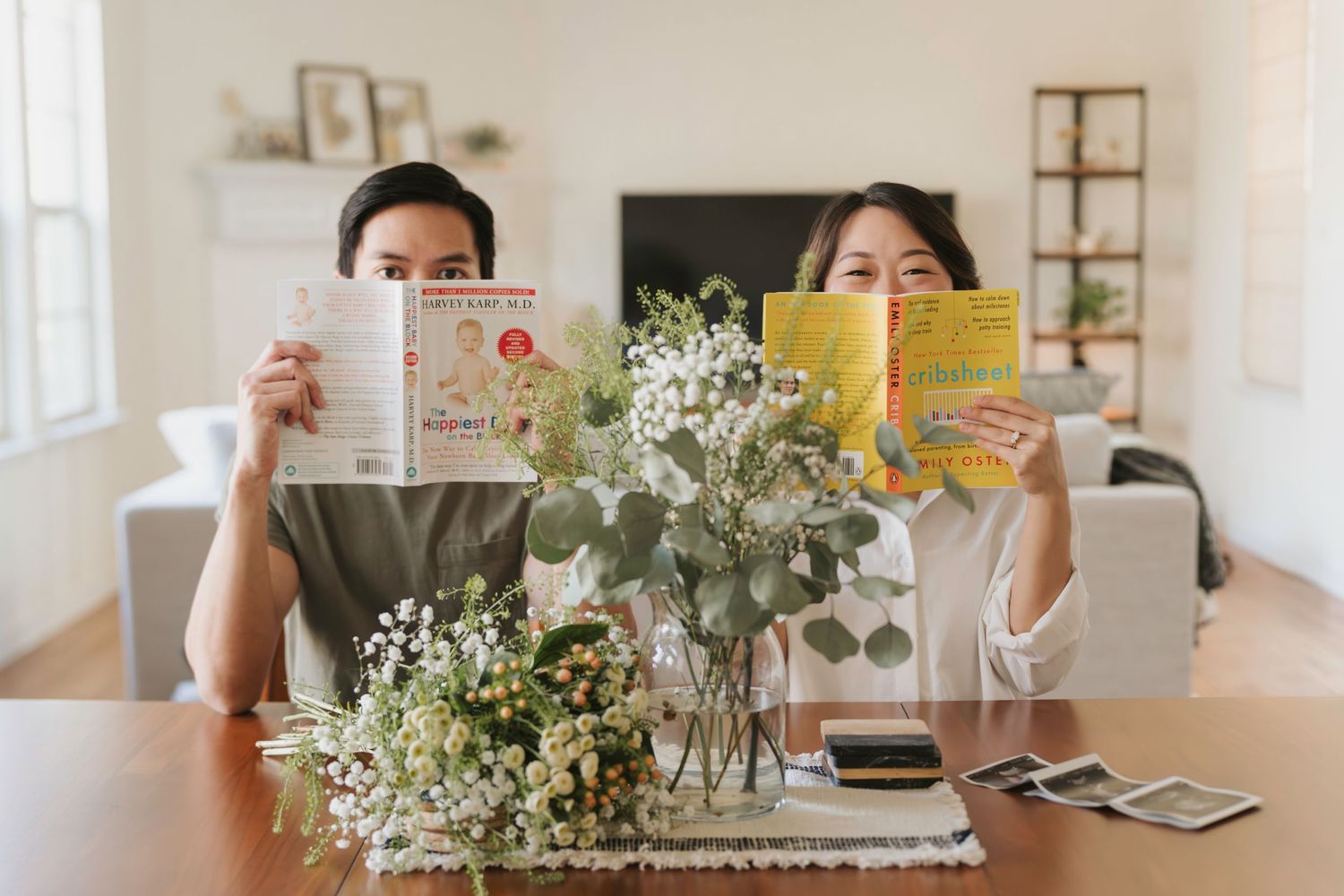 Two people holding books up at a table with a white floral centerpiece arrangement in glass vases.