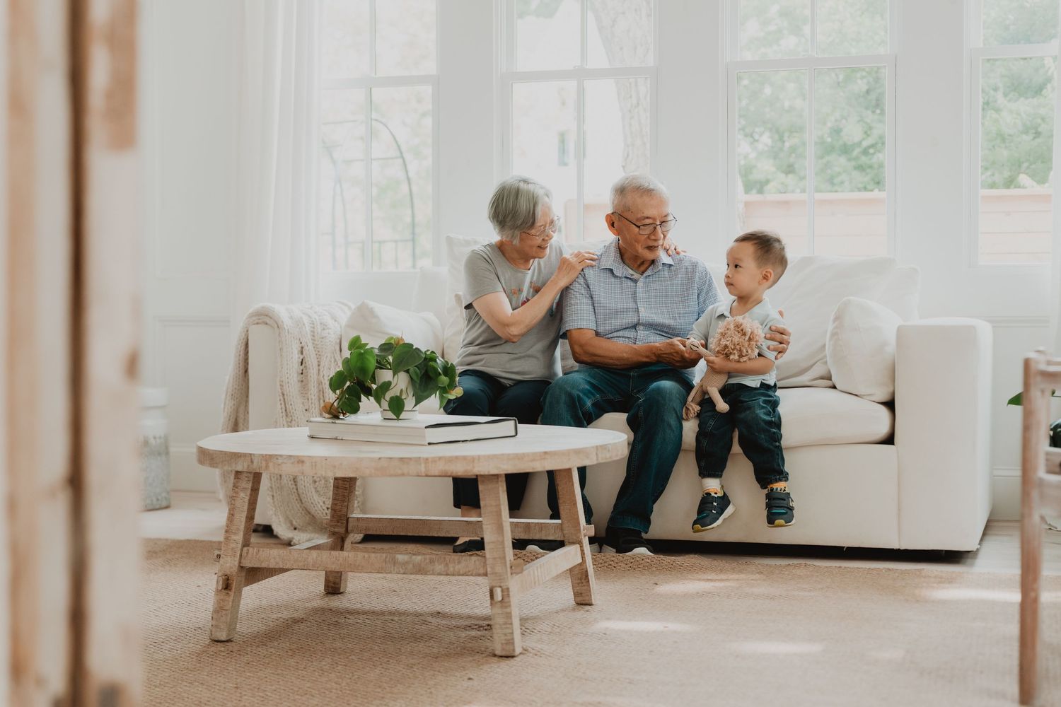 Elderly couple and young child sitting together on white couch in bright sunlit living room with wooden coffee table.