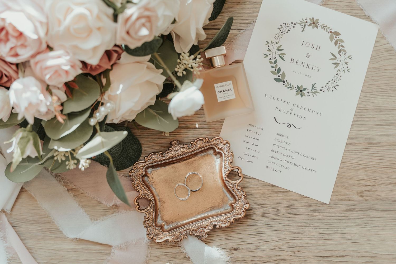 Wedding invitation with delicate floral wreath design alongside pink roses and vintage-style frame on wooden surface.
