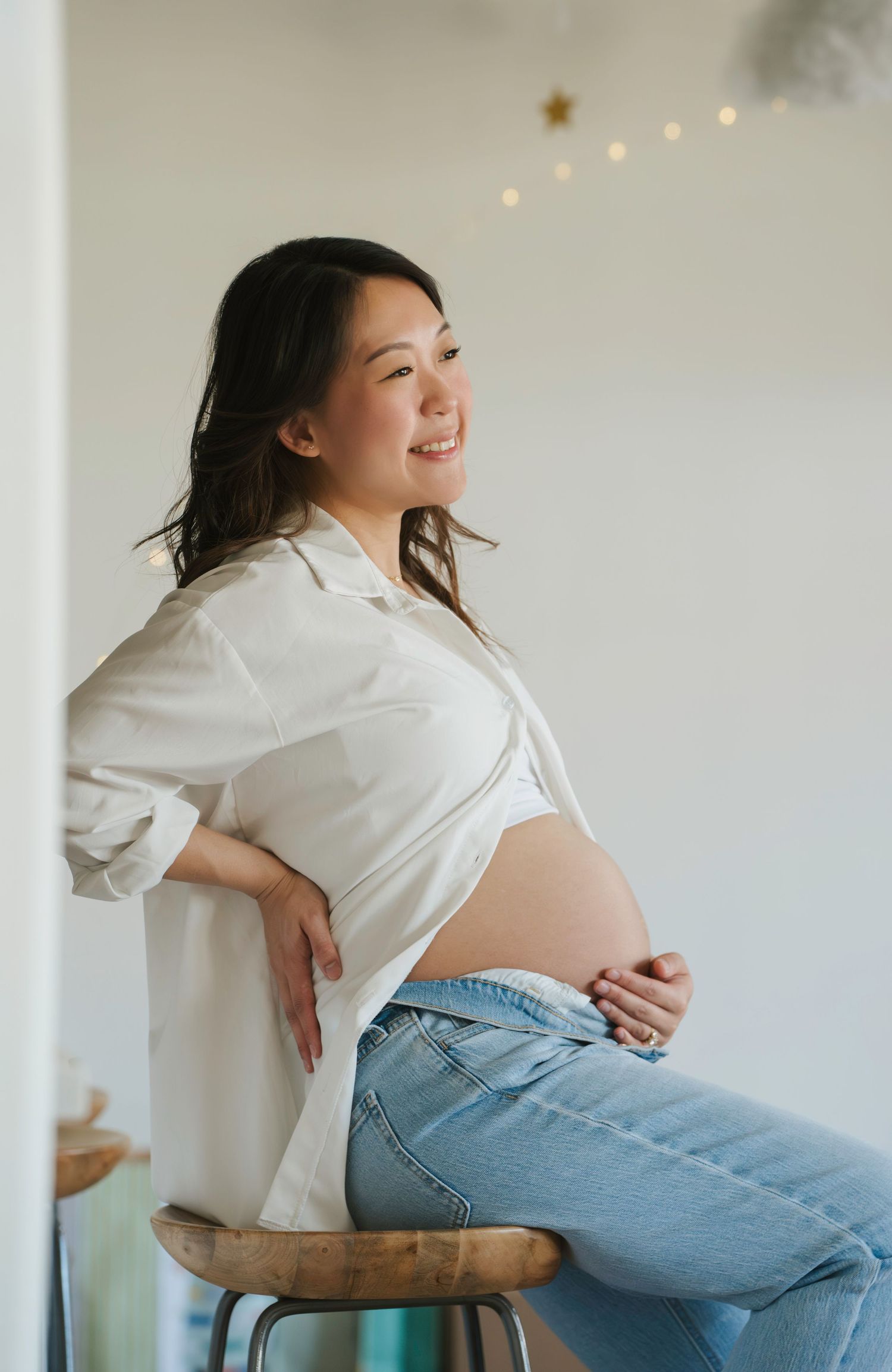 A pregnant woman in a white shirt and jeans sits casually on a stool while smiling and holding her belly.