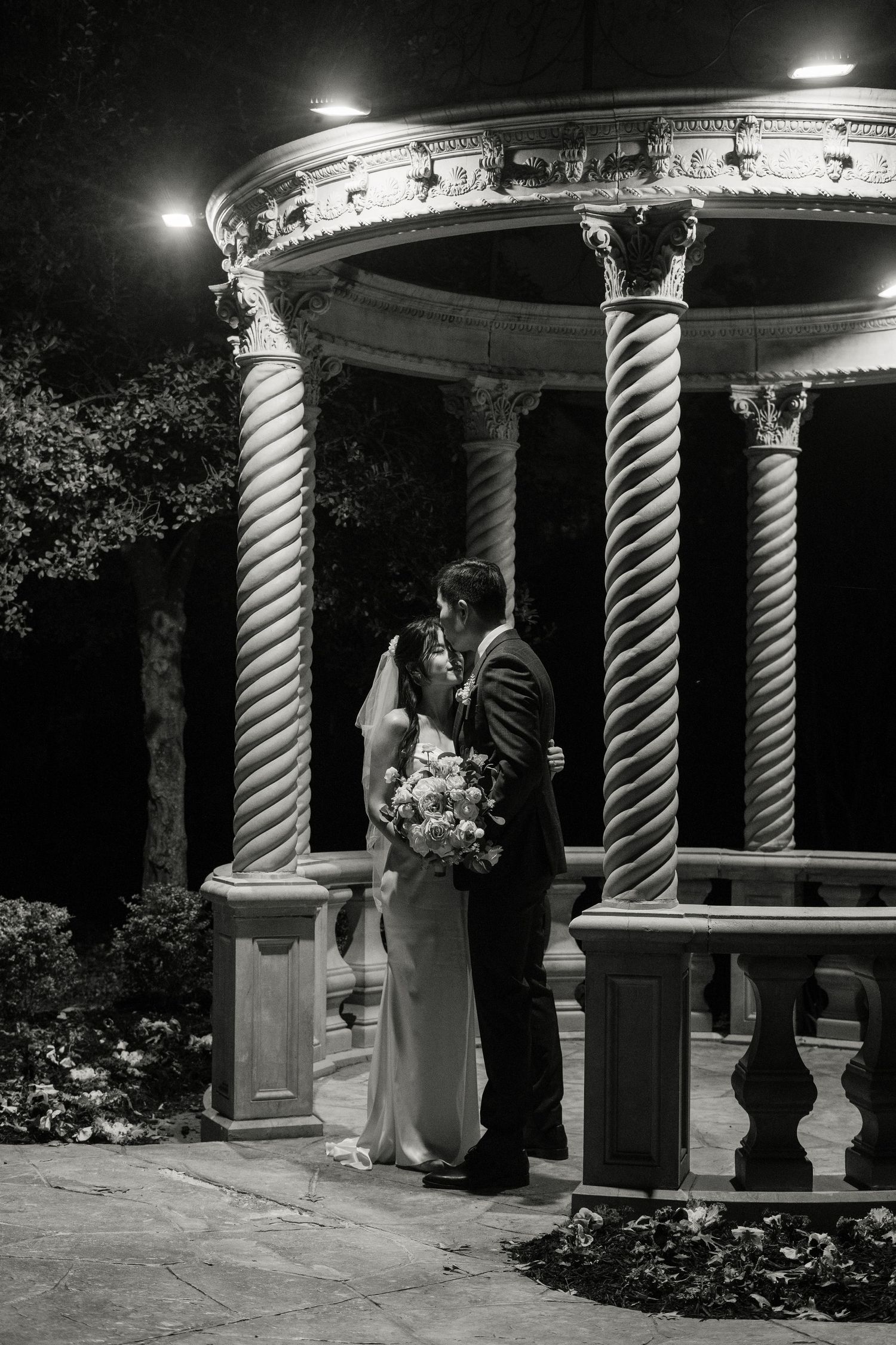 A romantic wedding couple shares a moment under an ornate gazebo with spiral columns illuminated at night.