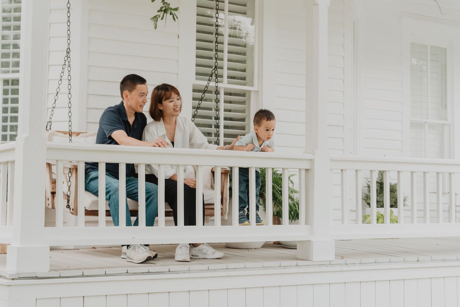 Three people sit together on a white porch swing at a charming farmhouse-style home with white columns and shutters.