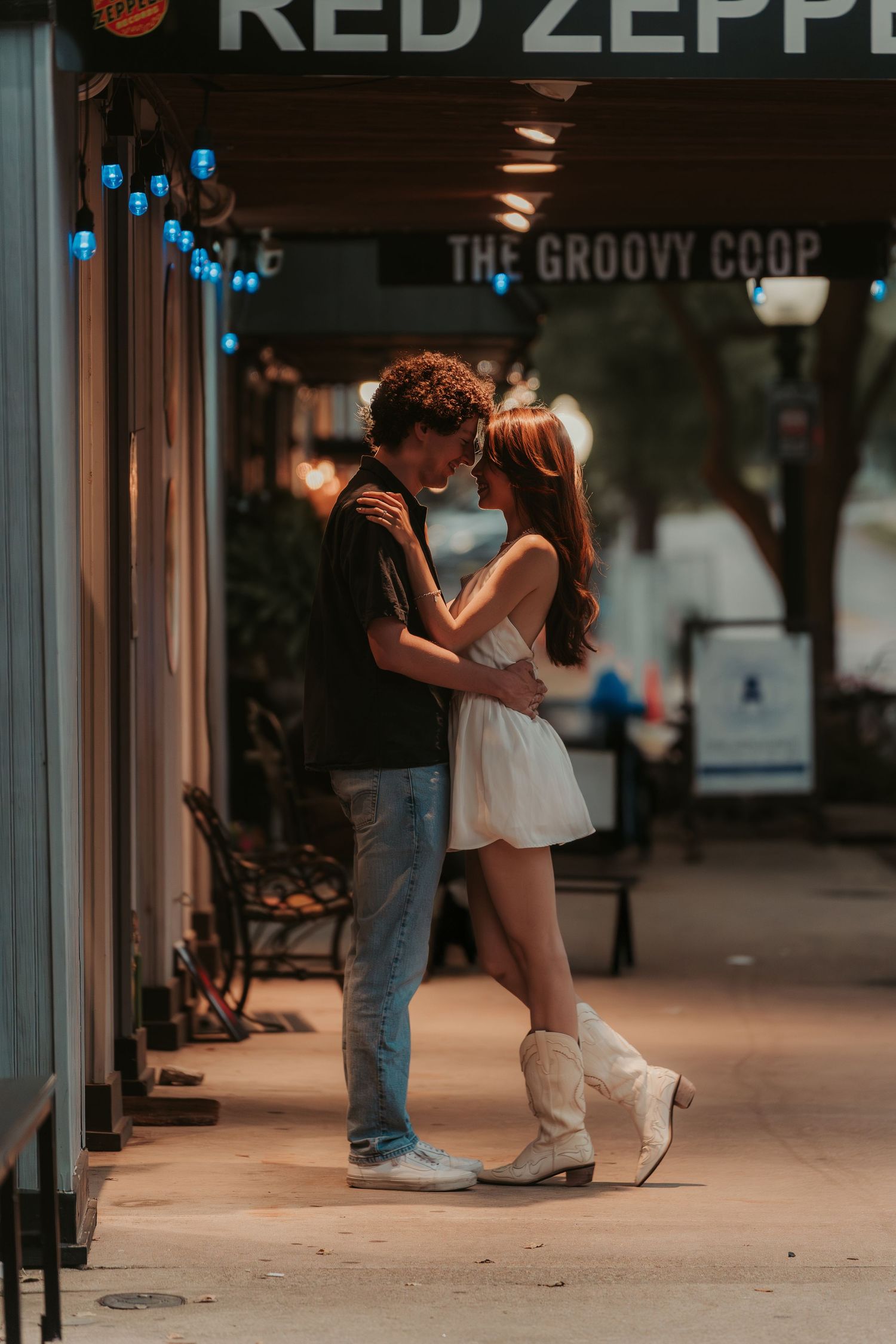 A romantic couple embraces under glowing blue lights outside a storefront with vintage signage at dusk.