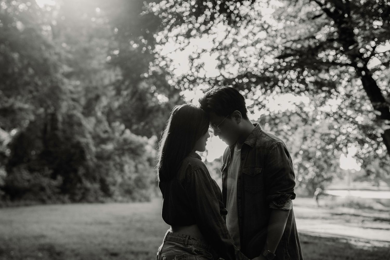 A romantic couple stands close together in a serene park setting with dreamy backlit sunlight filtering through trees.