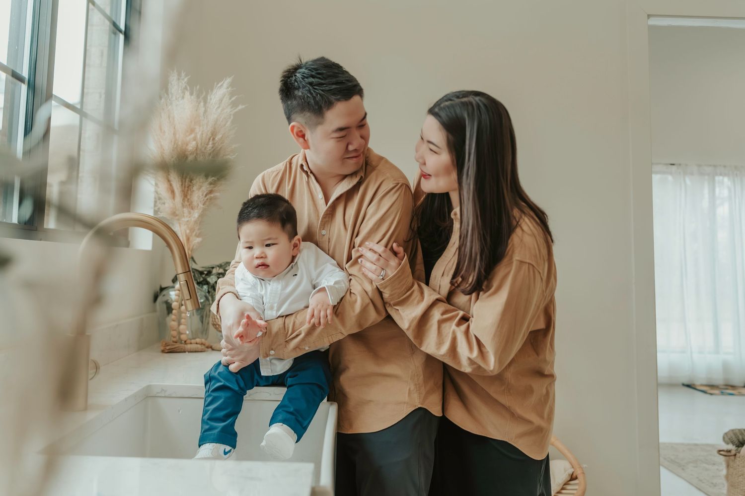 A family wearing matching beige sweaters poses together in their bright sunlit living room with pampas grass decor.