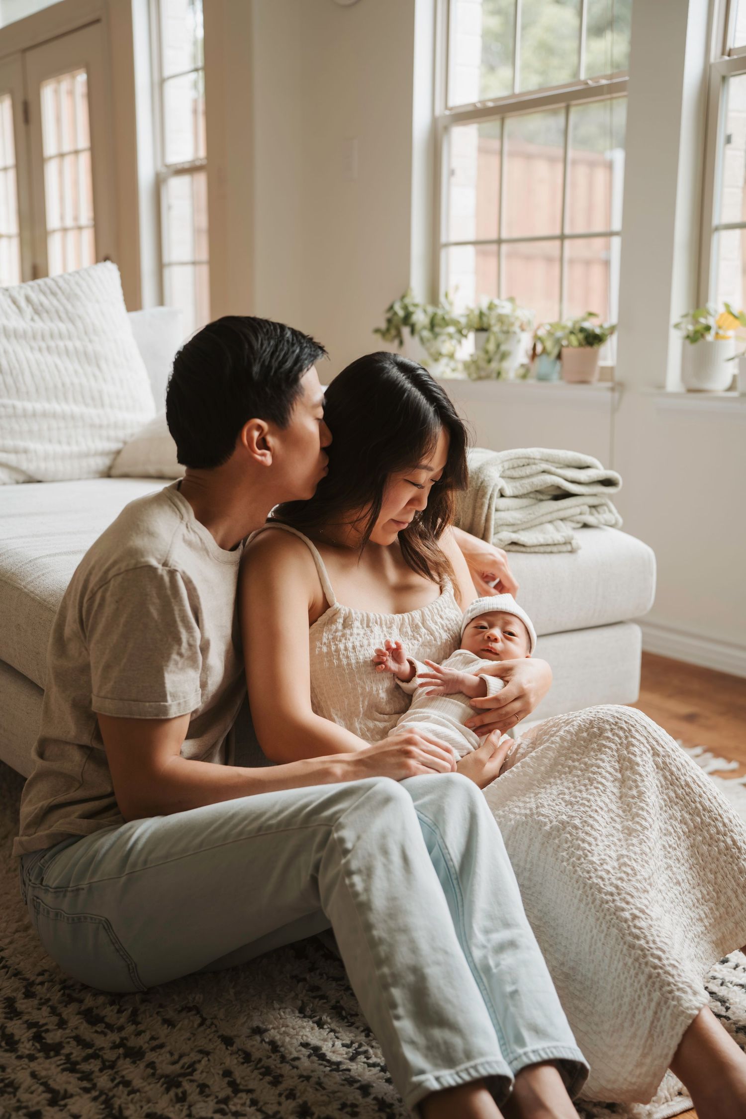 Parents cuddle with their newborn baby on a white couch in a sunlit living room with plants by the windows.