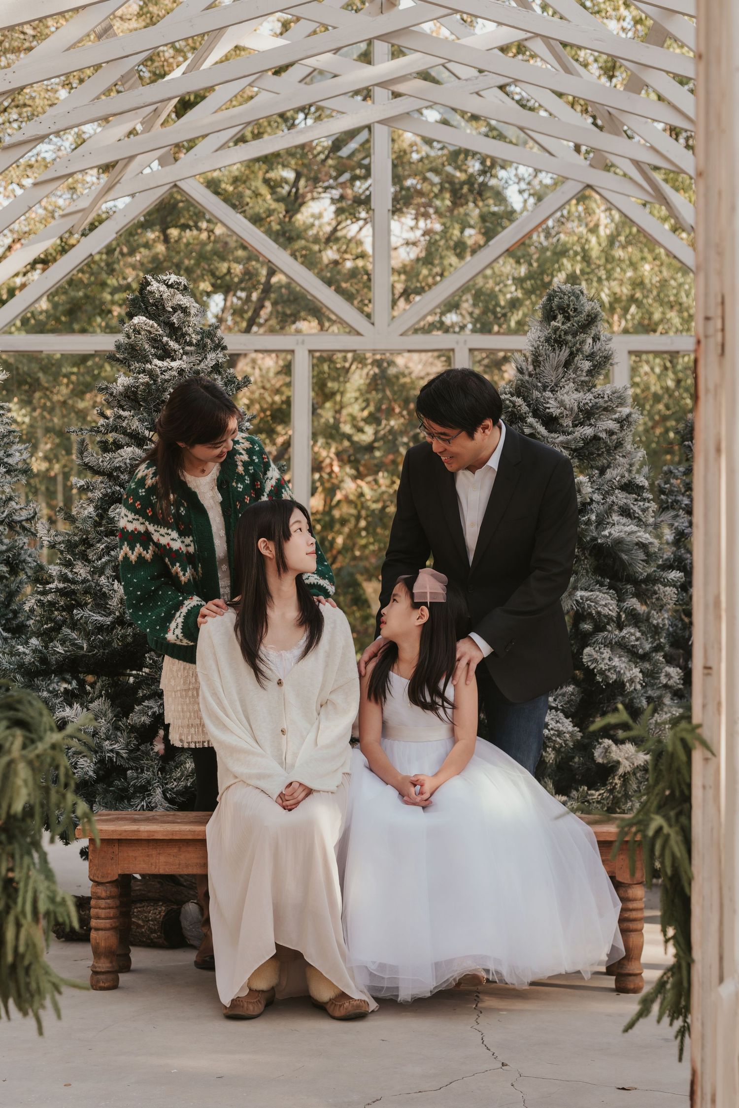 Wedding party sits casually on wooden bench in a bright greenhouse structure decorated with evergreen plants.